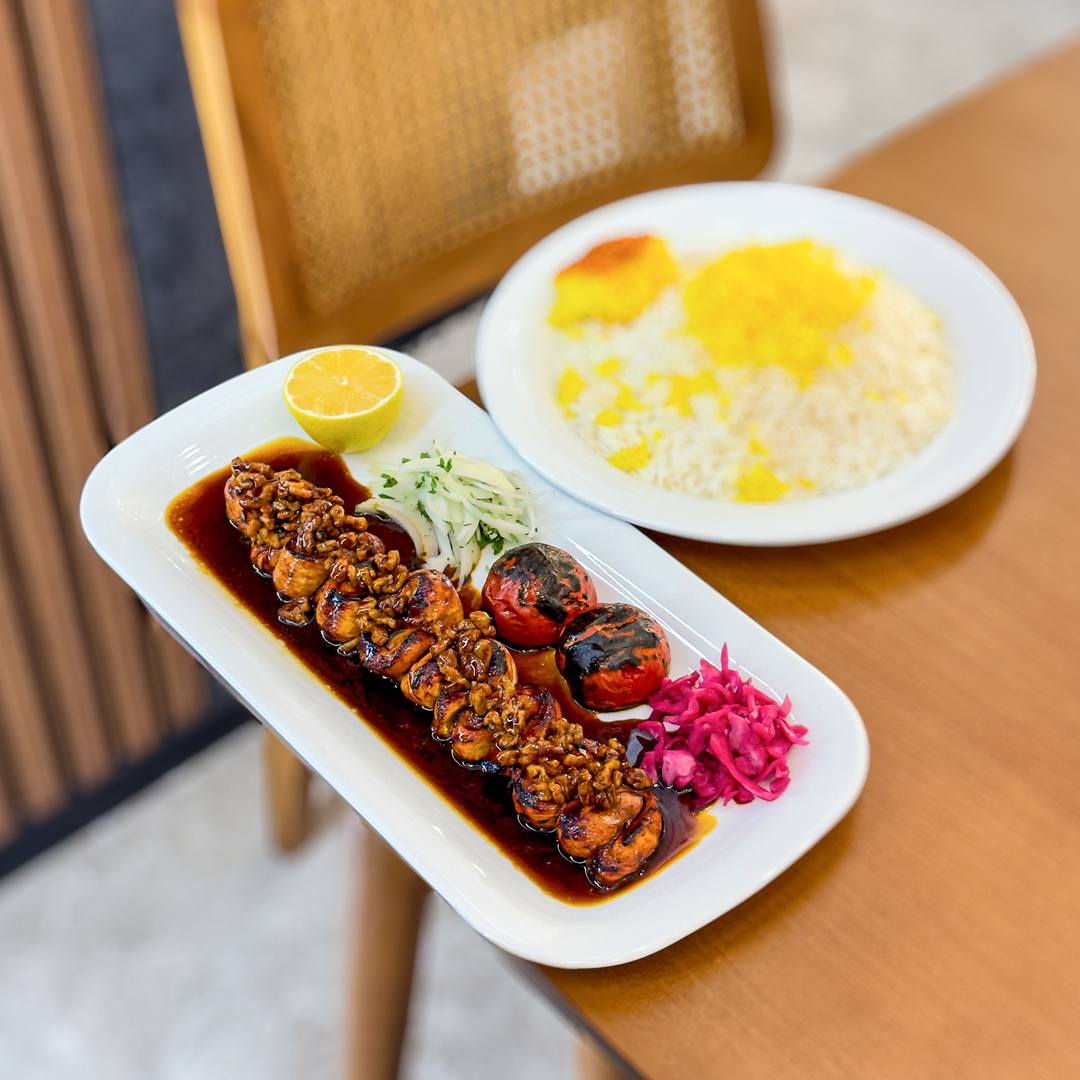 A low foot bridge hovers over reflective water. Above it is a dramatic tree whose leaves are bright orange and red. The leaves are falling slowly toward the water.