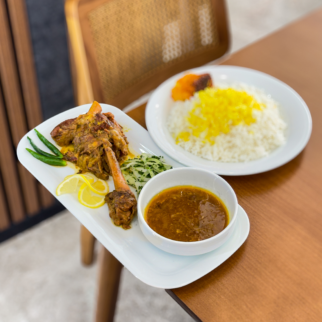 A medieval stone castle is shown at sunrise, the sky a mellow blue with trees and hills in the distance. There is a light frost on the ground, and small lights illuminating the grounds. There are lights glowing from within the few windows scattered across the facade and turret wings with capped roofs on each end of the peaked-roof structure.