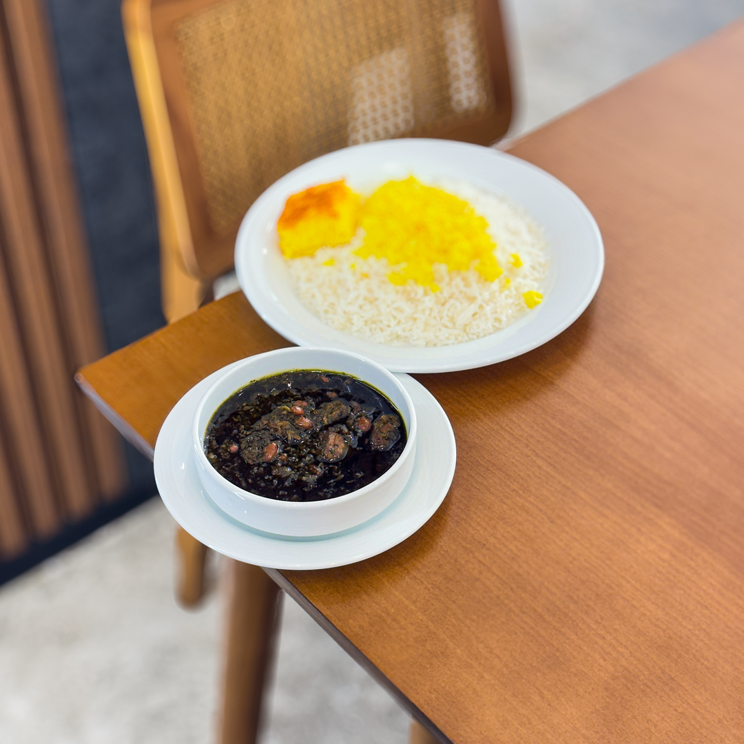 A medieval stone castle is shown at sunrise, the sky a mellow blue with trees and hills in the distance. There is a light frost on the ground, and small lights illuminating the grounds. There are lights glowing from within the few windows scattered across the facade and turret wings with capped roofs on each end of the peaked-roof structure.